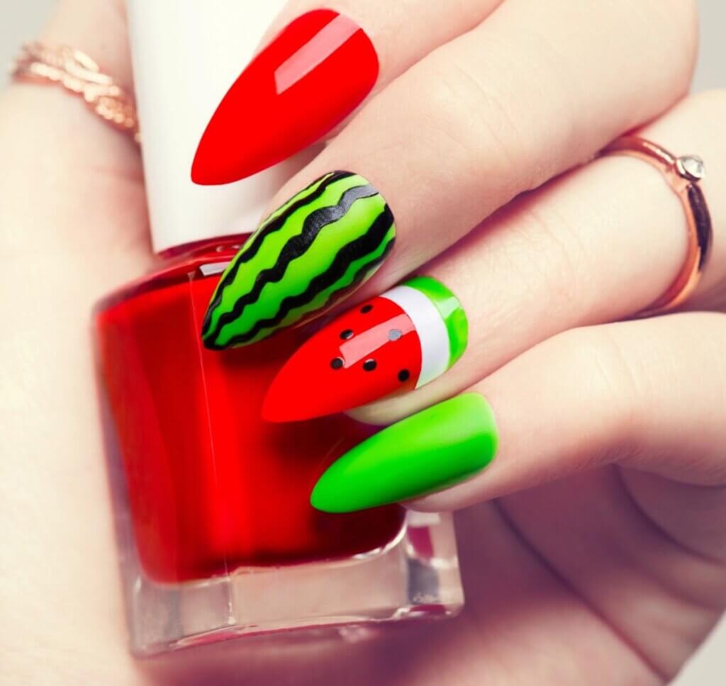 Trendy Summer Nails shown on a image featuring a hand with long nails painted with watermelon and a bottle of nail polish